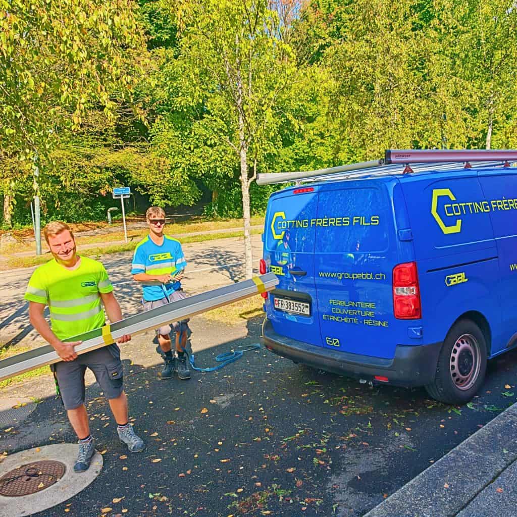 Deux ouvriers chargent une pièce métallique dans une camionnette bleue.